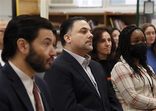 people sit while listening to a speaker (out of frame). The focus of the image on a man in the middle of the frame.